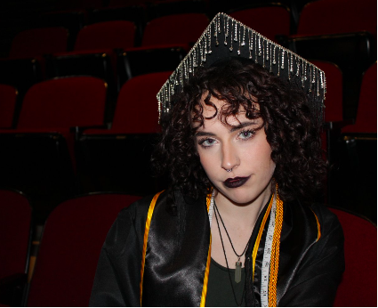 carolan, a girl with dark curly hair sitting in a theatre wearing graduation garb