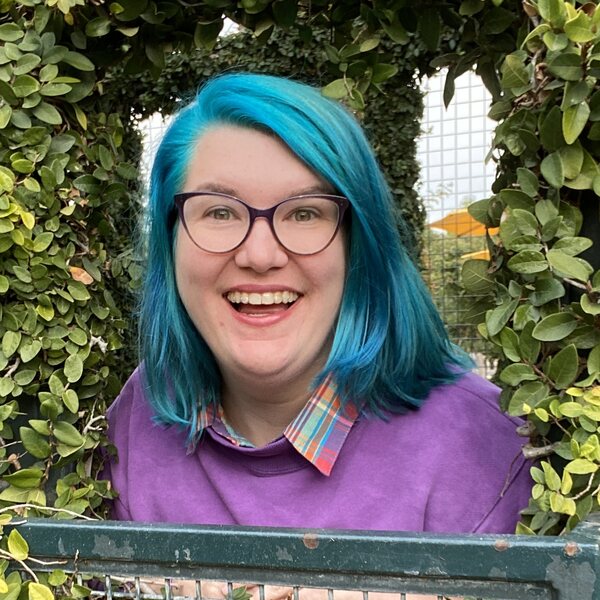 a white woman with bright blue hair and purple glasses smiles at the camera, surrounded by bushes