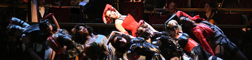 actor spotlighted in red corset with ensemble cast in black corsets in shadow around them