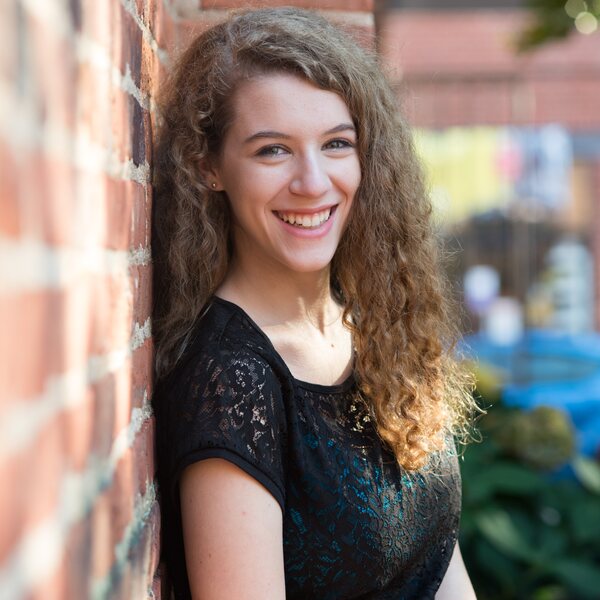 A smiling young woman with curly hair sitting against a brick wall