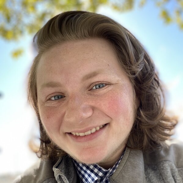Apollo, a White, young, transgender person looks at the camera sith his head tilted slightly to the RiGht. He is smiling, with bLe eyes and freckles. His blond hair is shoulder length. He is wearing a blue and white Checkered shirt and a grey vest.