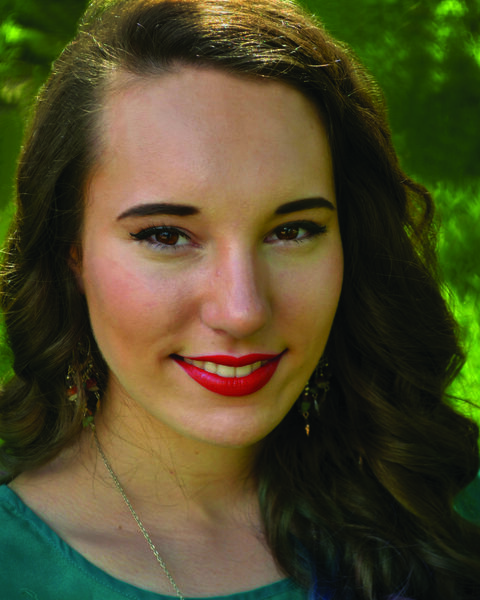 Jessica Lynn Hightower's Headshot. Close up of a woman with long, curled, brown hair and a teal shirt. The background is blurry green. 