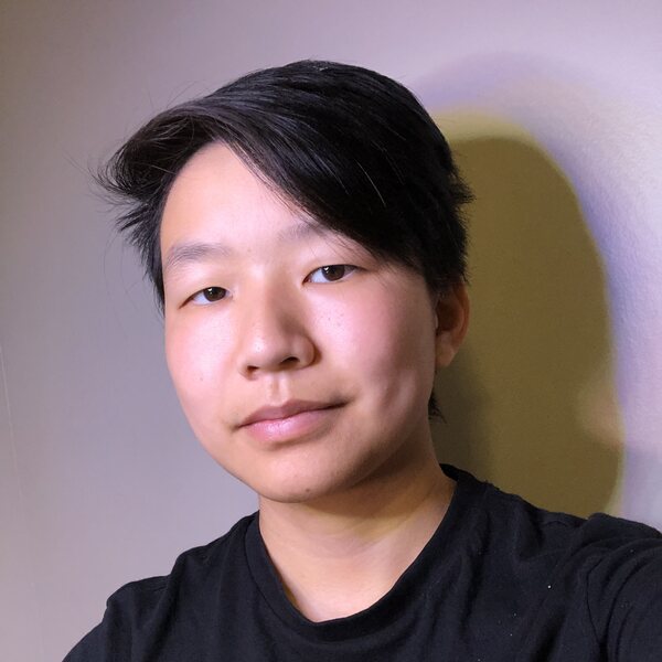 an asian american person in a black shirt with short dark hair, dark eyes, and tan skin. they have been photographed from the shoulders up against a grey background and are highlighted in a cool toned front light. they give a small smile toward the camera. 
