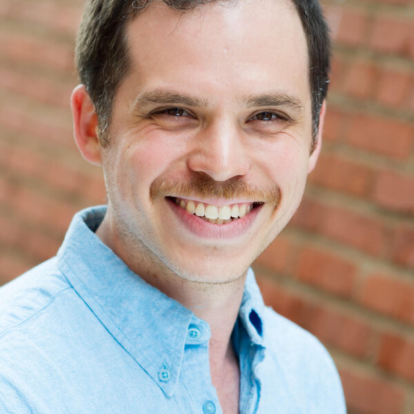headshot of eric swartz aka el chelito in front of a red brick wall. he has brown hair and eyes, a red mustache, and is smiling. he is wearing a pale blue shirt.