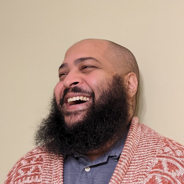 A smiling brown-skinned man with a bushy black beard looks off to the left of the frame. He wears a red cardigan and a lavender polo.
