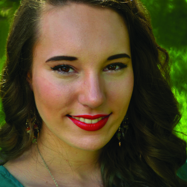Jessica Lynn Hightower's Headshot. Close up of a woman with long, curled, brown hair and a teal shirt. The background is blurry green. 